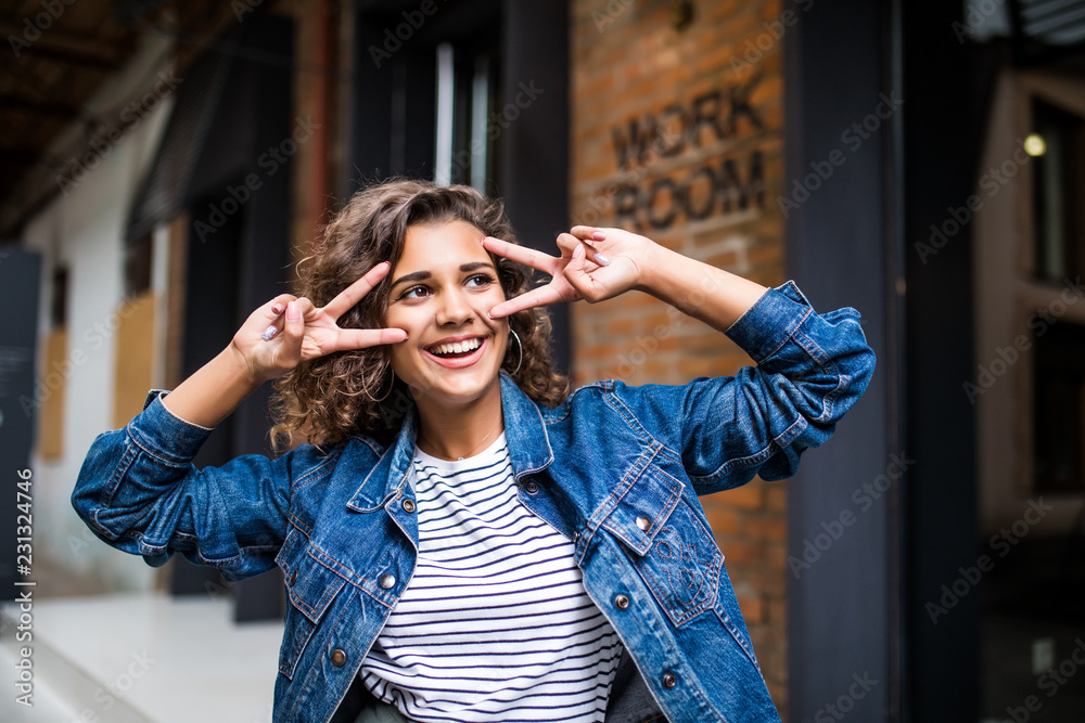 Young latin curly girl wearing a jeans jacket with peace gesture standing on a brick background on a street.