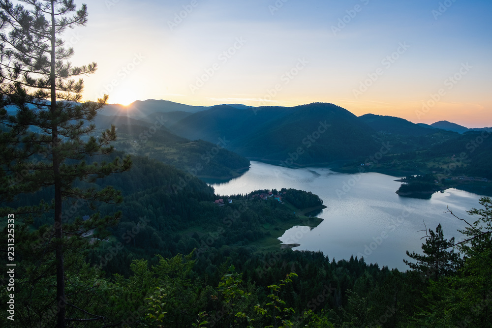 Lake among mountains with pine tree in the front during sunset. The sun almost desappeared in the mountains and rays of light lit the slopes of hills Lake among mountains with pine tree in the front during sunset. The sun almost desappeared in the mountains and rays of light lit the slopes of hills