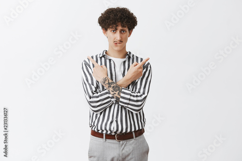 Help me make decision. Weak and insecure concerned stylish caucasian guy with curly hairstyle and moustache crossing hands on chest pointing left and right biting lip being unsure which way right