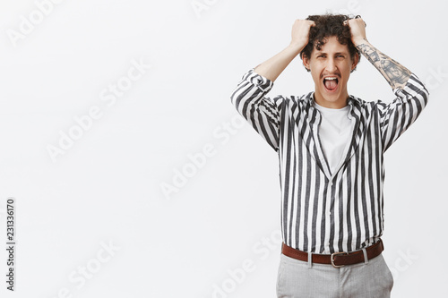 Studio shot of depressed young guy with moustache in striped shirt losing temper screaming out loud pulling hair out of head being fed up and pissed off by stupid boss posing over gray background