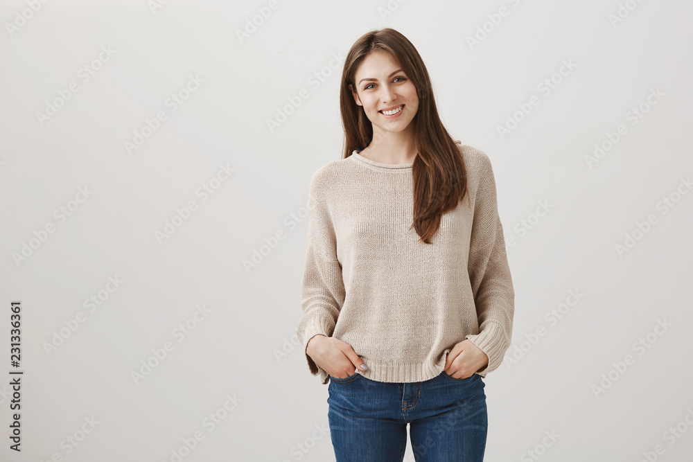 Friendly newbie meets with new teammates. Indoor shot of shy attractive female in casual pullover standing with hands in pockets and tilting head while smiling at camera, talking to stranger Friendly newbie meets with new teammates. Indoor shot of shy attractive female in casual pullover standing with hands in pockets and tilting head while smiling at camera, talking to stranger