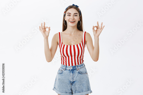 Friendly beautiful young woman in trendy pin-up striped top and shorts showing okay gesture and smiling assured and happy liking awesome design of flat friend make posing against gray wall