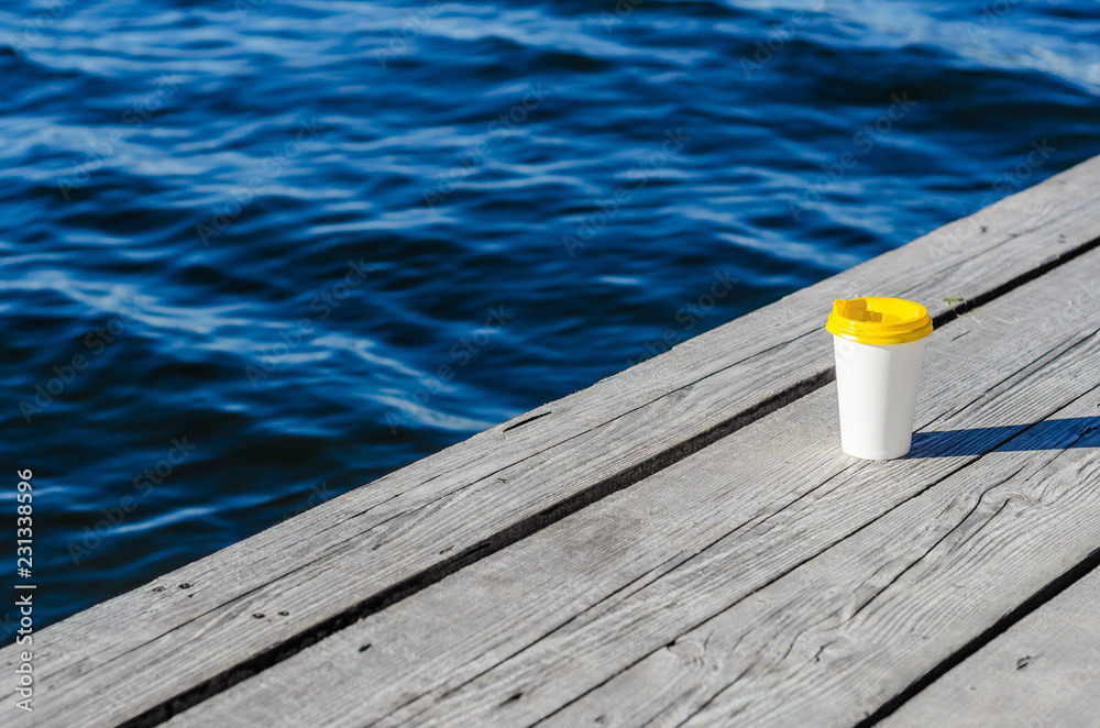White paper cup with a yellow plastic lid stands on the boards on the shore of the lake
