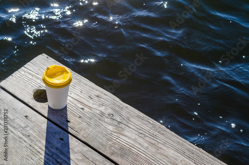 White paper cup with a yellow plastic lid stands on the boards on the shore of the lake