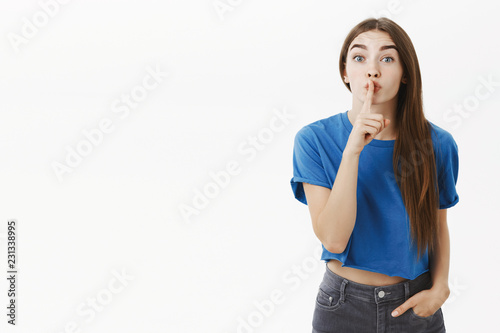 Excited energized woman asking keep secret safe not telling anoying posing with shush gesture against grey wall showing shh sign with index finger over mouth wanting hide information over grey wall photo