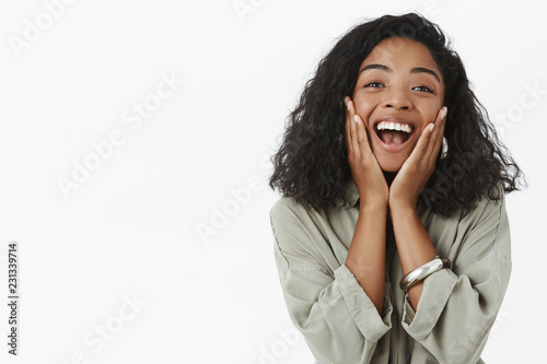 Waist-up shot of excited happy charming African American girlfriend on vacation touching cheeks from amazement and joy yelling yeah and smiling joyfully posing charmed and delighted over gray wall