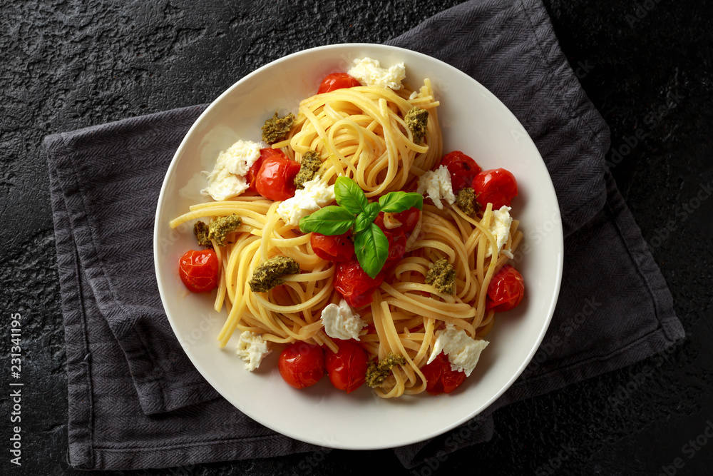 Pasta with green pesto sauce, roasted cherry tomatoes and mozzarella cheese in white plate on dark rustic background