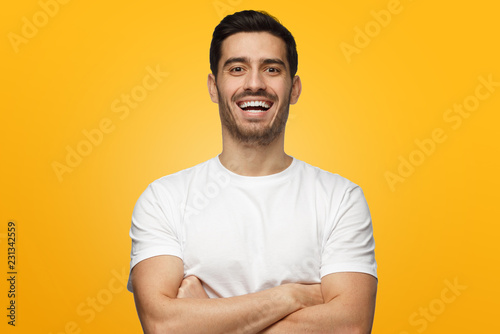 Young handsome man isolated on yellow background, with arms crossed, showing open friendly happy friendly smile