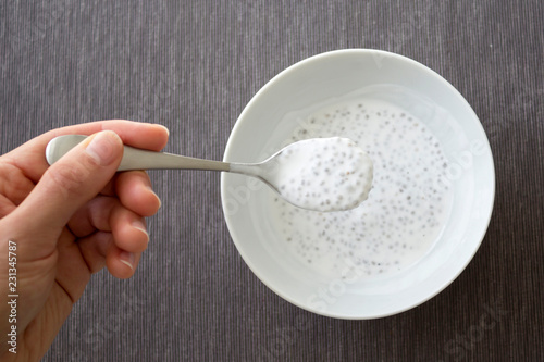Hand holding the spoon of the plate of chia seeds with coconut milk, grey tablecloth background, top view