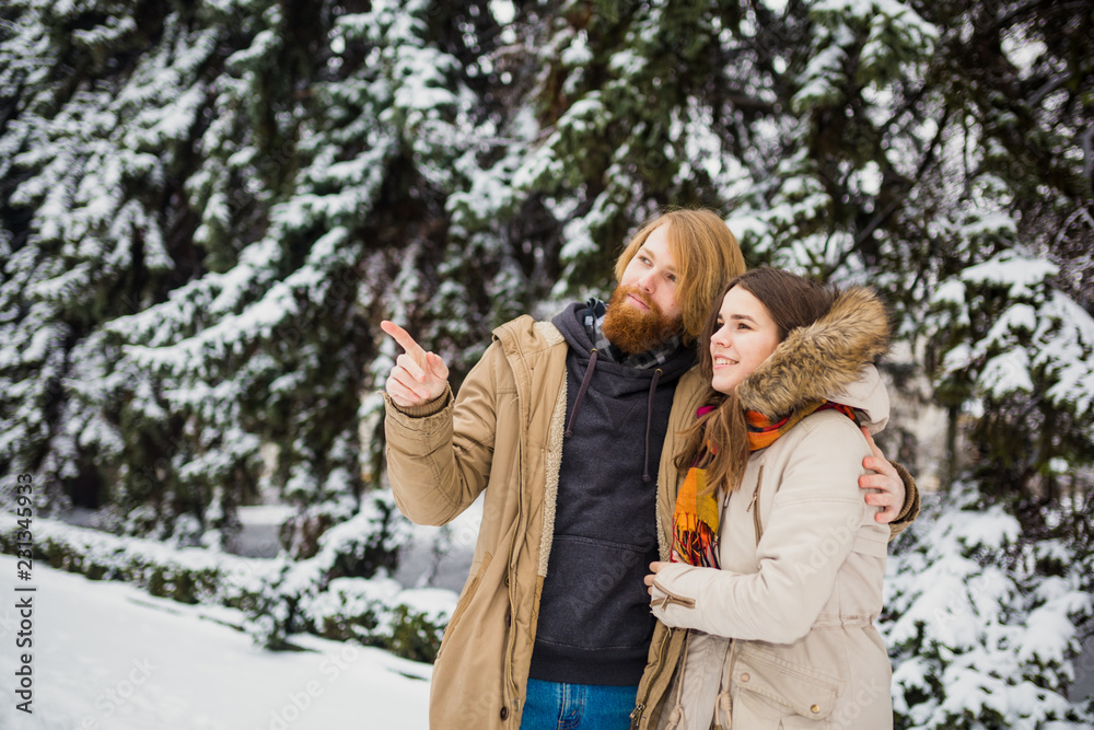 Young Caucasian boy and girl in love heterosexual couple have a date outdoors in a winter park on the background of a snowy conifer standing in an embrace. Man hugging woman valentines day Young Caucasian boy and girl in love heterosexual couple have a date outdoors in a winter park on the background of a snowy conifer standing in an embrace. Man hugging woman valentines day