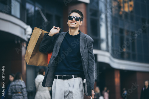 Serious stylish young man with sunglasses walking in urban street and enjoying Black Friday shopping in trendy stores in city