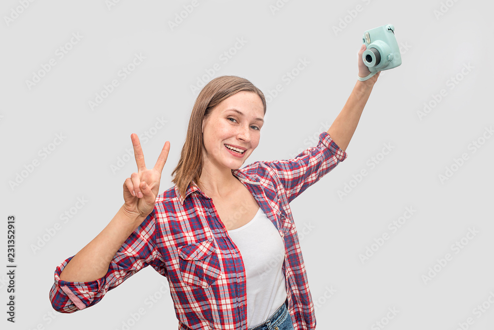Obraz premium Young woman holding blue camera in hand and stretch arm up. She poses and smiles. Woman looks straight. She shows piece symbol with fingers. Isolated on grey background.