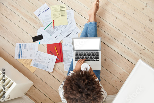 Top view of darrk skinned female deals with bills and taxes, works on laptop computer with white mock up screen, drinks takeaway coffe, poses on wooden floor at domestic setting, makes report photo