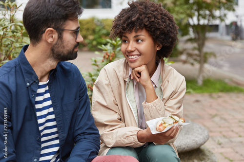 Multiethnic couple have stroll outdoor, rest in city, eat sandwich, look positively at each other, discuss future plans. Groupmates eat fast food after classes. City life and leisure time concept