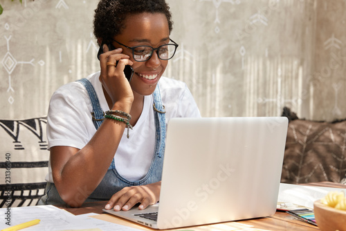 Stylish African American woman calls on smart phone looks in laptop computer, updates software, has phone conversation, checks banking balance for making transaction, has piercing, dressed casually