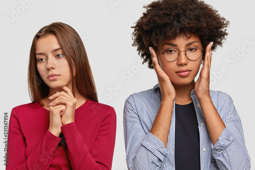 Thoughtful Caucasian lady looks pensively with sad expression aside, turns back from her Afro girlfriend after quarrel, have bad relatioships, isolated over white wall. Dark skinned girl covers ears photo