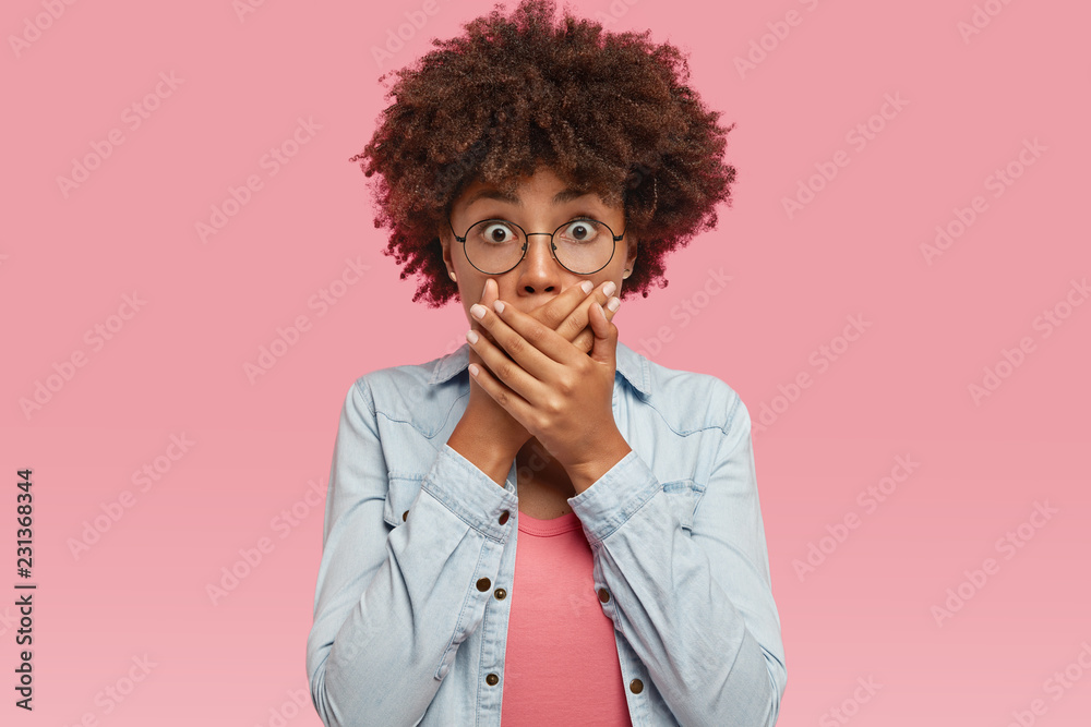 Shot of pleasant looking young ethnic female covers mouth with both palms, tries not spread rumors, being shocked with gossiping, stares through round spectacles, stands against pink background. Shot of pleasant looking young ethnic female covers mouth with both palms, tries not spread rumors, being shocked with gossiping, stares through round spectacles, stands against pink background.