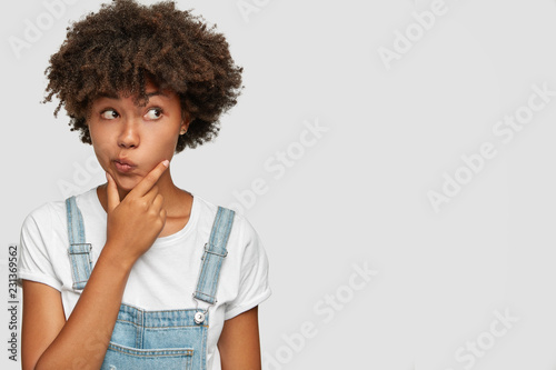 Candid shot of confused clueless woman holds chin, purses lips with hesitation, looks aside, wears fashionable clothes, poses against white background with copy space for your promotional text