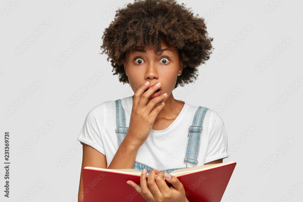 Surprised Frightened Black Girl Covers Mouth From Amazement Doesnt Believe In Shocking News Holds Textbook Keeps Eyes Popped Out Poses Against White Background Facial Expressions Concept Stock Photo Adobe Stock