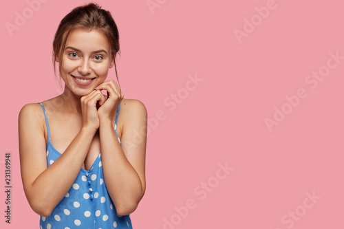 Pretty young lady with positive expression, keeps both hands near chin, smile...