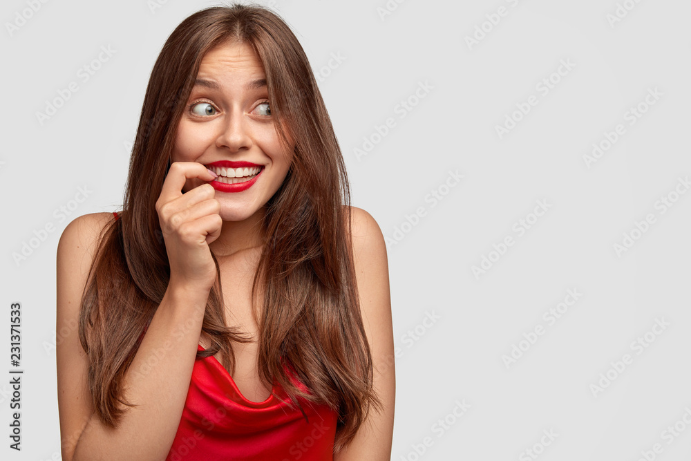 Positive European woman with mysterious happy expression, keeps index finger near mouth, smiles broadly, wears red lipstick, dressed in elegant dress, models over white wall with free space on right Positive European woman with mysterious happy expression, keeps index finger near mouth, smiles broadly, wears red lipstick, dressed in elegant dress, models over white wall with free space on right