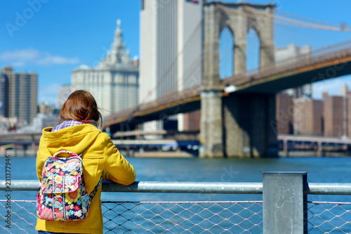 Happy young woman tourist sightseeing by Brooklyn Bridge, New York City, at sunny spring day. Female traveler enjoying view of downtown Manhattan.