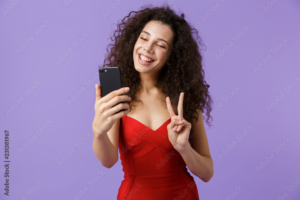 Image of charming woman 20s wearing red dress using black smartphone, standing isolated over violet background Image of charming woman 20s wearing red dress using black smartphone, standing isolated over violet background