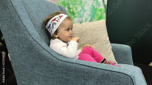 Little baby girl sitting in armchair looking away, smiling and pointing a finger at something. Toddler raptly watching TV and vocalising with emotions. Child interest to cartoon or moving pictures photo