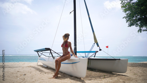 Wide shot of beautiful brunette woman in red swimwear, she sits on the edge of the boat located on the beach © South House Studio