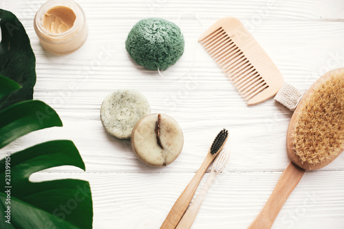 Zero waste flat lay. Natural solid shampoo bar, bamboo toothbrushes, wooden brush, deodorant cream and konjaku sponge on white wood with green monstera leaves. Eco products plastic free