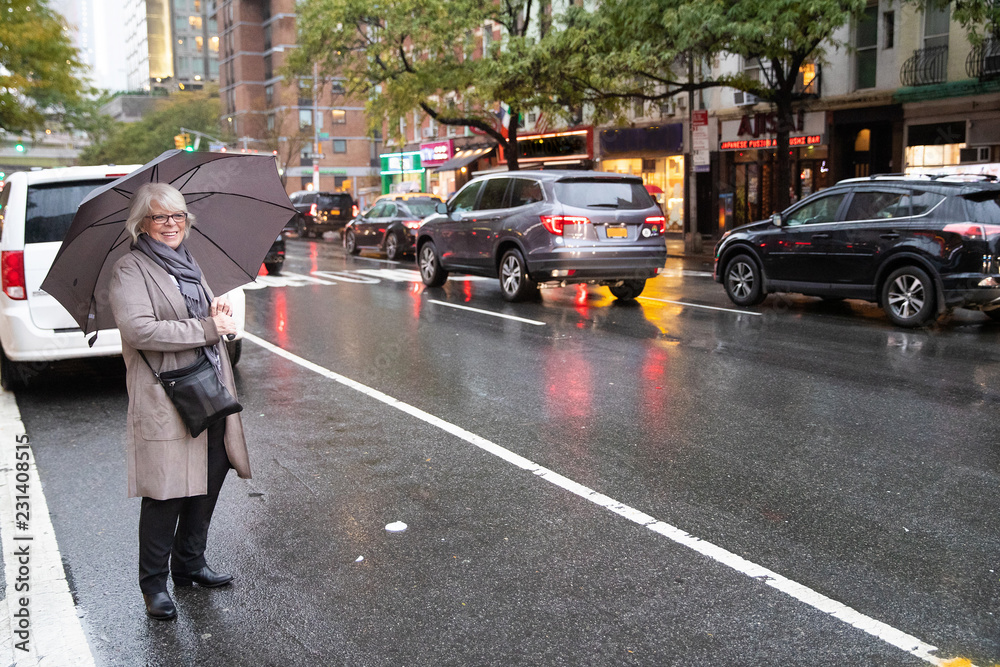 Fototapeta premium Mature senior woman standing on busy New York street under umbrella in the rain waiting for ride share car service