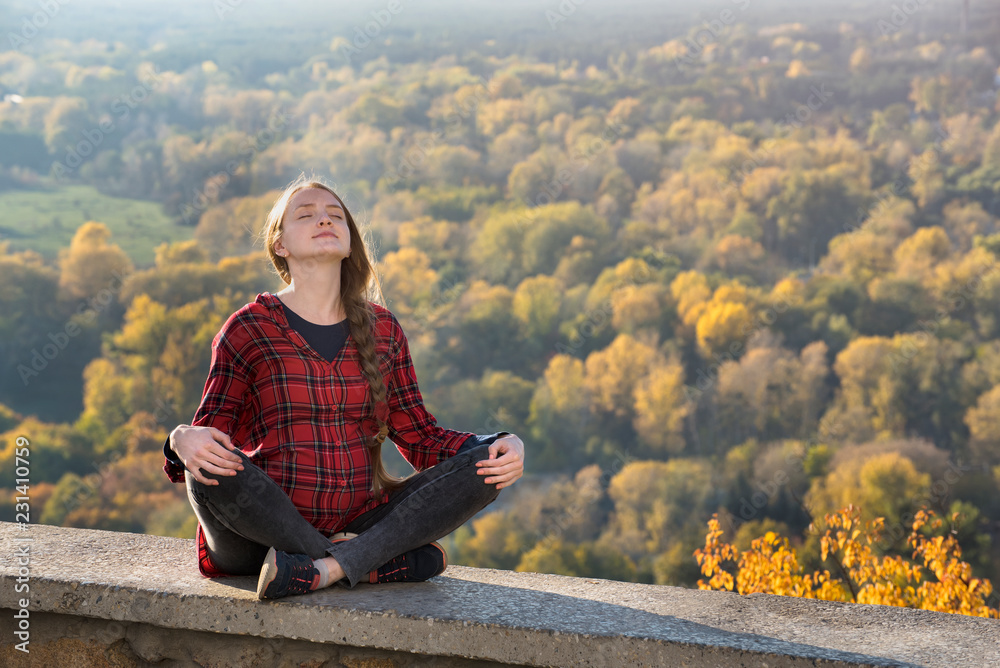 Pregnant woman sits on a hill with her eyes closed. Meditation. Autumn forest on the background