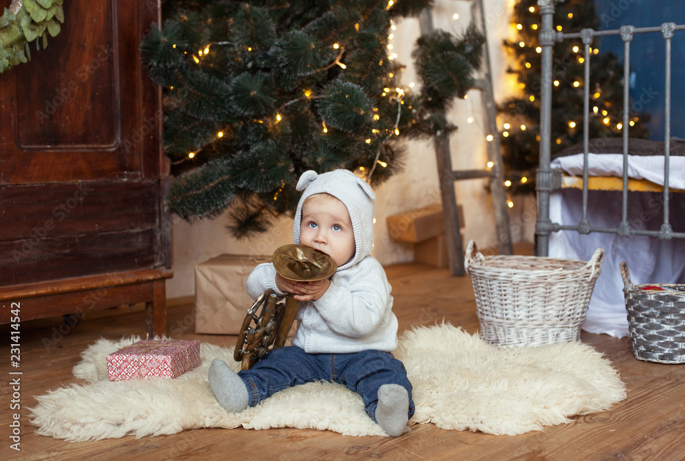 Obraz premium A little boy is sitting with a gift and a copper pipe on the background of the Christmas tree, a vintage cabinet and a basket in retro style. New Year, Christmas card in a Scandinavian interior