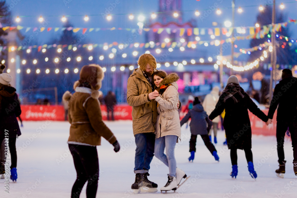 Naklejka premium Young couple in love Caucasian man with blond hair with long hair and beard and beautiful woman have fun, active date skating on ice scene in town square in winter on Christmas Eve