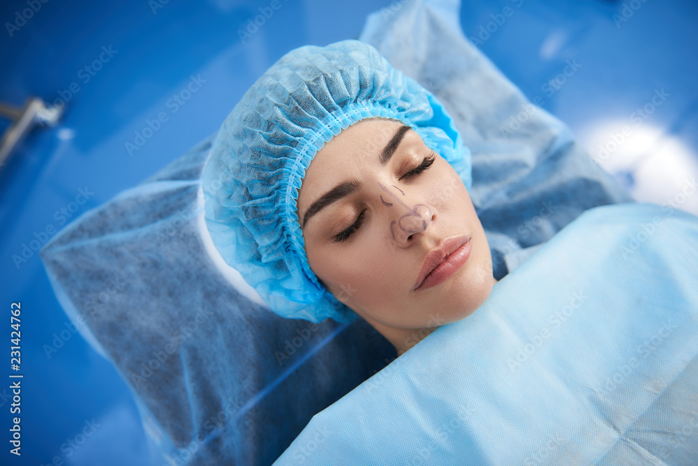 Calm pretty patient lying in the operation room with her eyes closed and having lines on her face before the plastic surgery Calm pretty patient lying in the operation room with her eyes closed and having lines on her face before the plastic surgery
