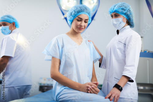 Kind doctor. Caring young doctor standing next to her patient and looking at her while putting one hand on her shoulder