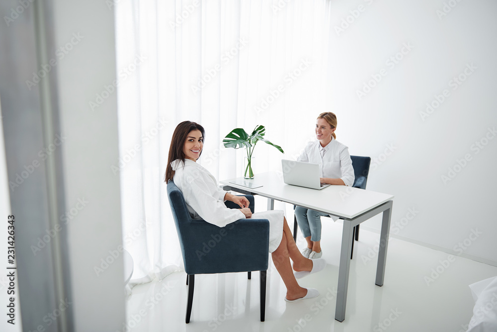Full length portrait of attractive lady in white bathrobe sitting at the table with beautician. They looking at camera and smiling Full length portrait of attractive lady in white bathrobe sitting at the table with beautician. They looking at camera and smiling