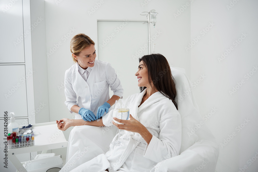 Fototapeta premium Portrait of charming woman sitting in armchair and holding glass of lemon water while doctor in sterile gloves checking IV infusion
