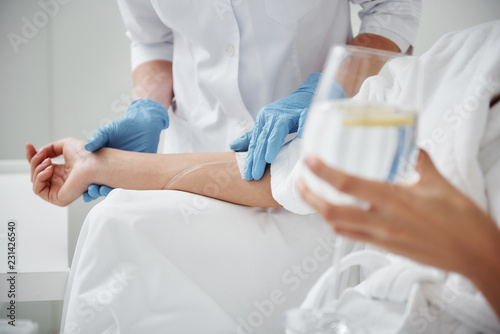 Close up of physician hand in sterile glove checking tube and needle for IV infusion on woman arm. Girl holding glass with lemon water photo