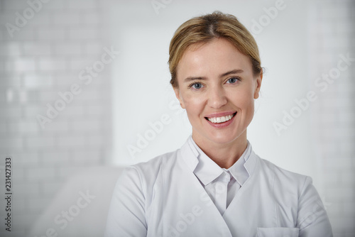 Welcome to our beauty salon. Portrait of young cosmetologist in white lab coat looking at camera with smile