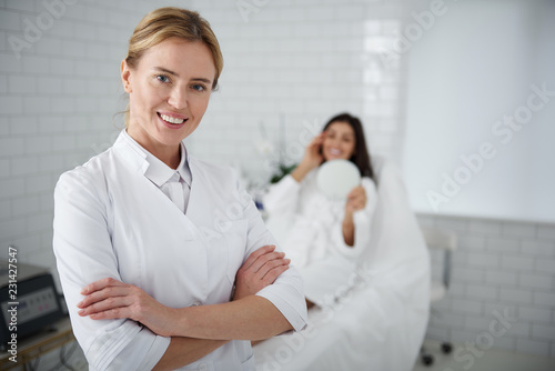 Waist up portrait of charming beautician in white lab coat crossing arms and looking at camera with smile. Young woman in bathrobe looking in the mirror on blurred background photo