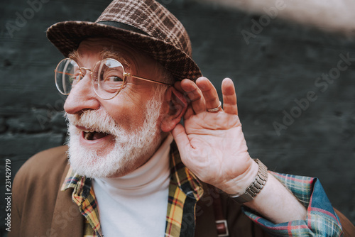 What. Close up portrait of stylish pensioner with playful glance holding palm near his ear and smiling