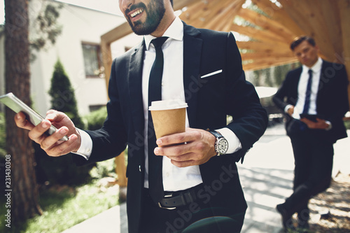 Young man in elegant suit standing outdoors and enjoying his coffee while looking at the screen of a modern device