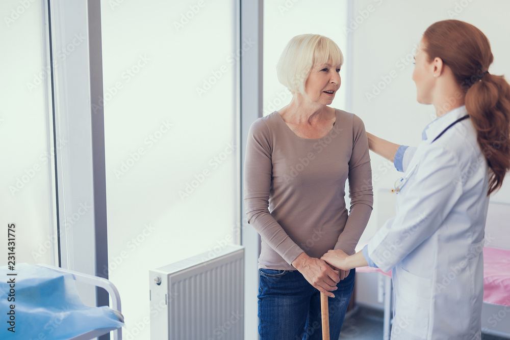 Friendly therapist is communicating with woman in office. They are standing jointly and practitioner is hugging patient with crutch. Copy space in left side Friendly therapist is communicating with woman in office. They are standing jointly and practitioner is hugging patient with crutch. Copy space in left side