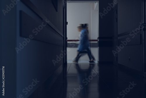 Focus on dark hallway of hospital. At its end patients and medical workers are walking. Copy space in left side
