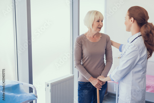 Friendly therapist is communicating with woman in office. They are standing jointly and practitioner is hugging patient with crutch. Copy space in left side