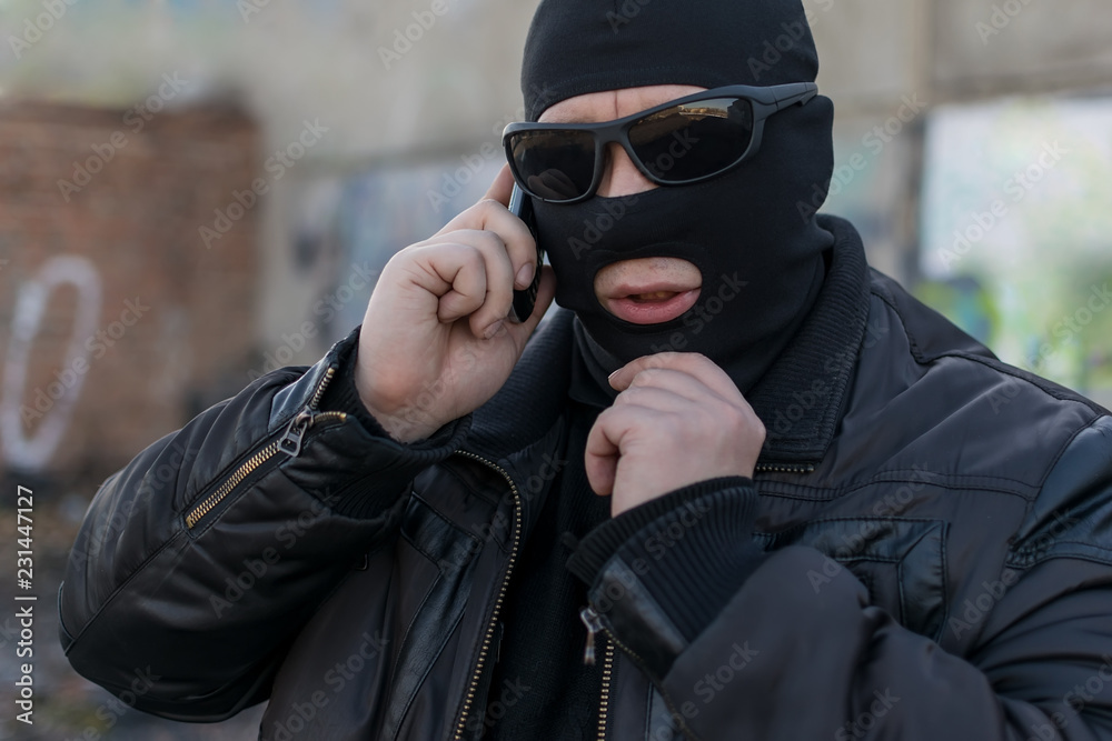 a man, a terrorist, a bandit in a black leather jacket and a mask talking  on the phone on the street near an abandoned building Photos | Adobe Stock