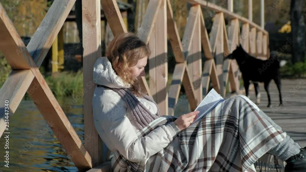 a woman on the bridge near the autumn river is reading a book a black dog are walking nearby