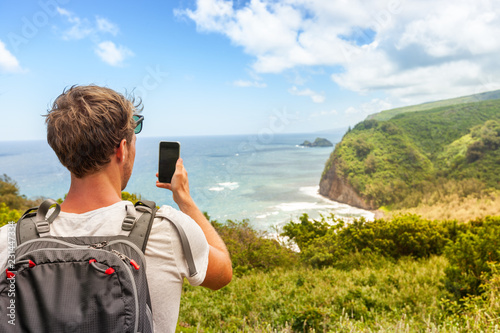 Travel tourist man in Hawaii beach USA vacation taking photo with mobile phone device of ocean landscape mountains background. Pololu Valley beach hike, Big Island, Hawaii.