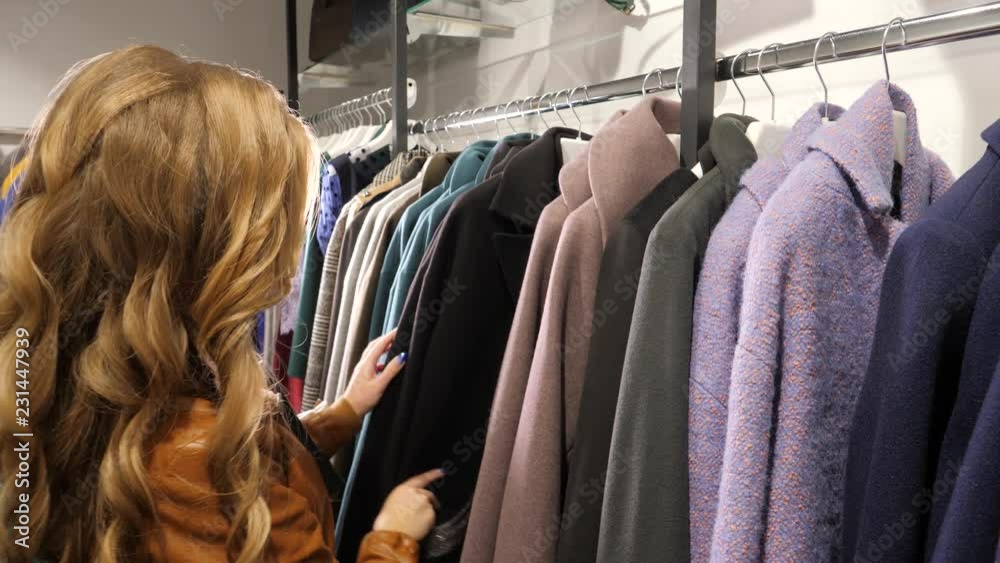 Young female shopping in a fashion boutique store watching coats and dresses on hangers Young female shopping in a fashion boutique store watching coats and dresses on hangers